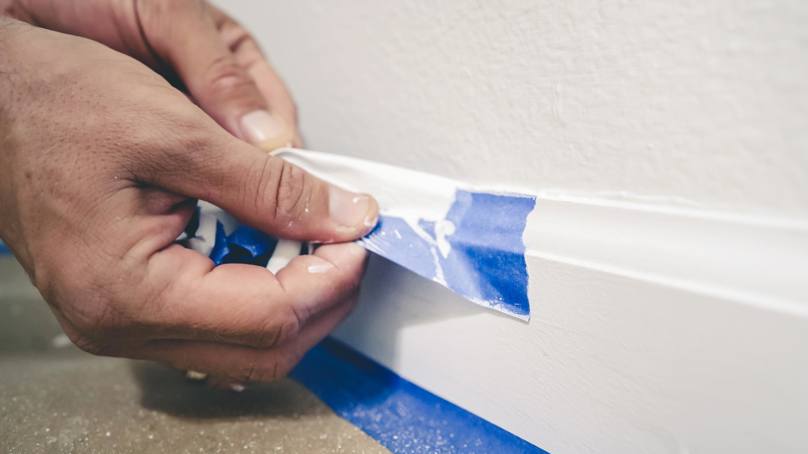 Person Removing Blue Masking Tape From Skirting Board After Painting