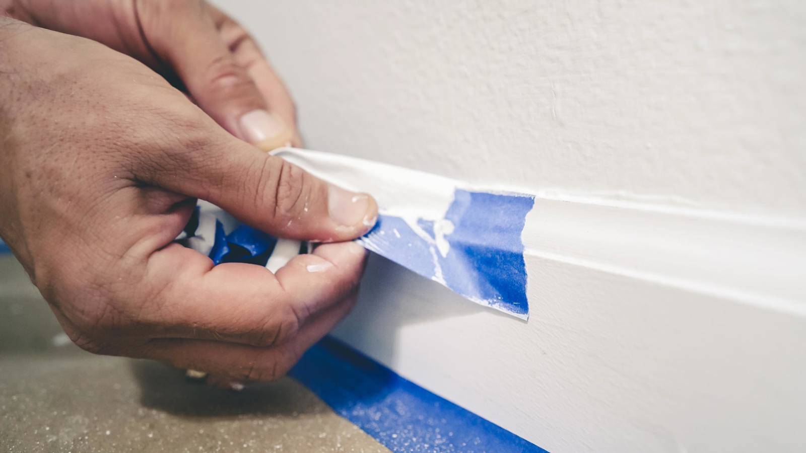 Person removing blue masking tape from skirting board after painting